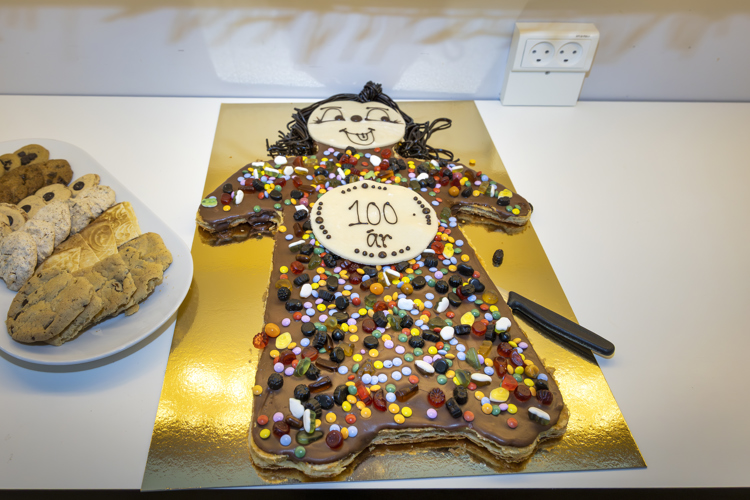 A large, colorful cake shaped like a person, decorated with candies and a "100 år" sign, alongside a plate of assorted cookies.