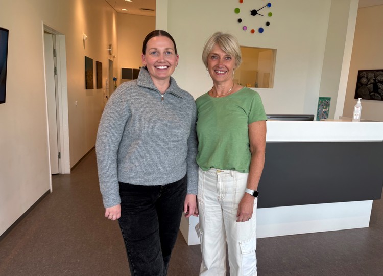 Two women stand smiling in a modern reception area. One wears a gray sweater and black pants, the other a green shirt and white pants.