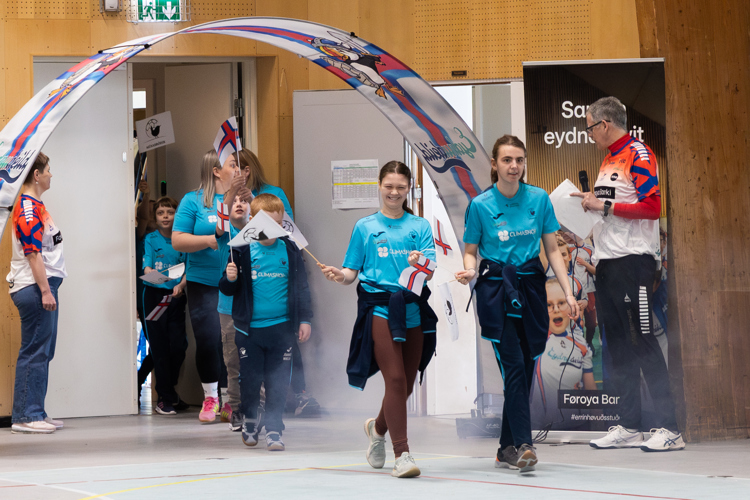 A group of children in blue shirts enters a gym, waving flags and smiling, with a banner in the background.