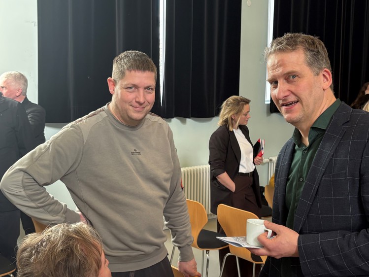 Two men are smiling and chatting in a room with black curtains. One holds a cup, while a woman stands in the background.