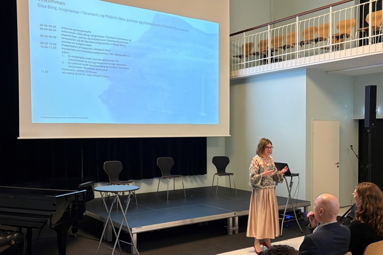 A woman speaks at a conference, with a presentation screen behind her. Chairs are arranged on stage, and attendees are seated in front.