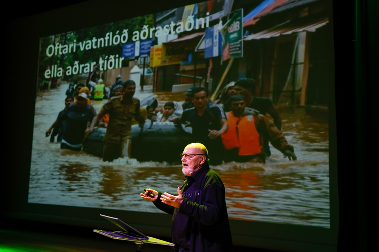 A speaker presents on stage with a flood rescue image projected behind, highlighting the impact of water disasters.