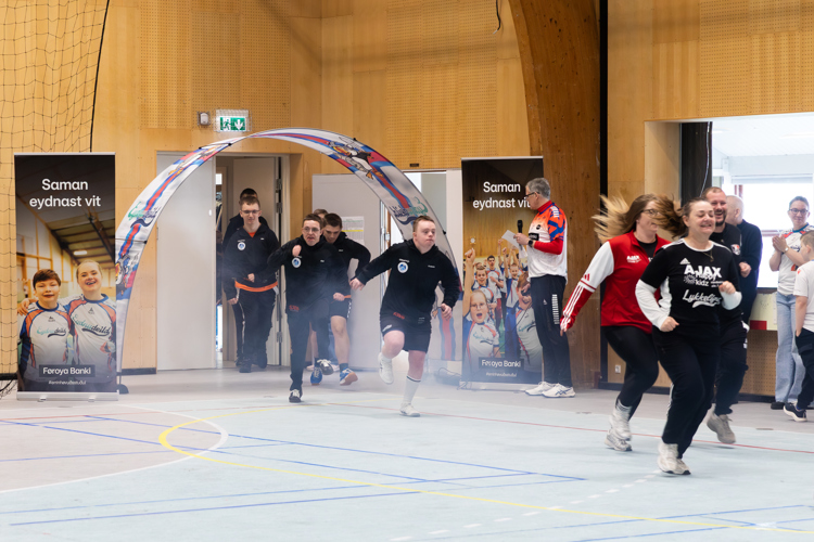 Participants are running into a sports hall through an arch, with banners in the background and an audience watching.