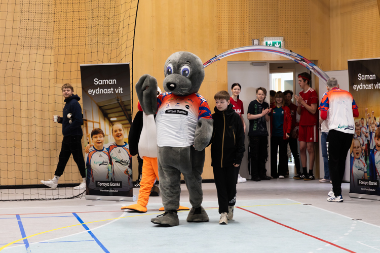 A mascot in a sports jersey waves while walking with children in a gym, with banners in the background.