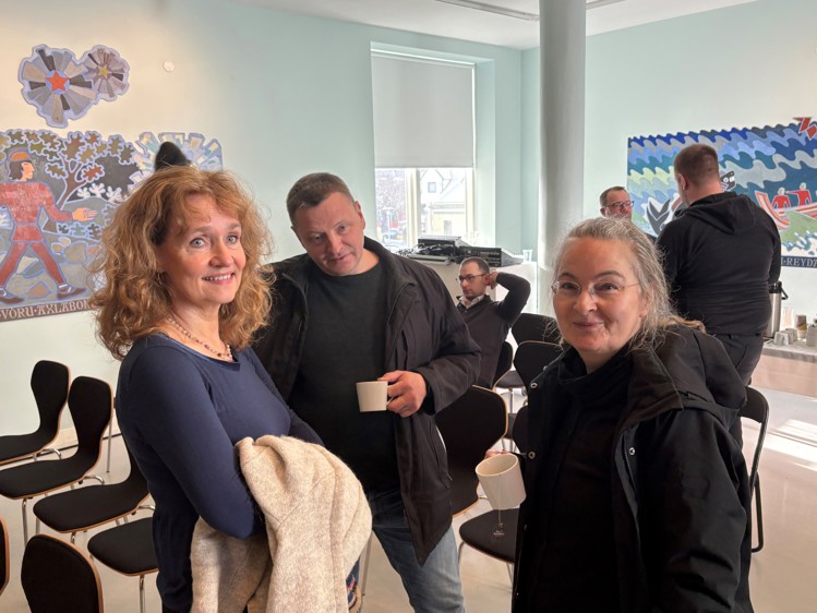 A group of people chatting indoors, with two women and a man in the foreground. Colorful artwork decorates the walls.