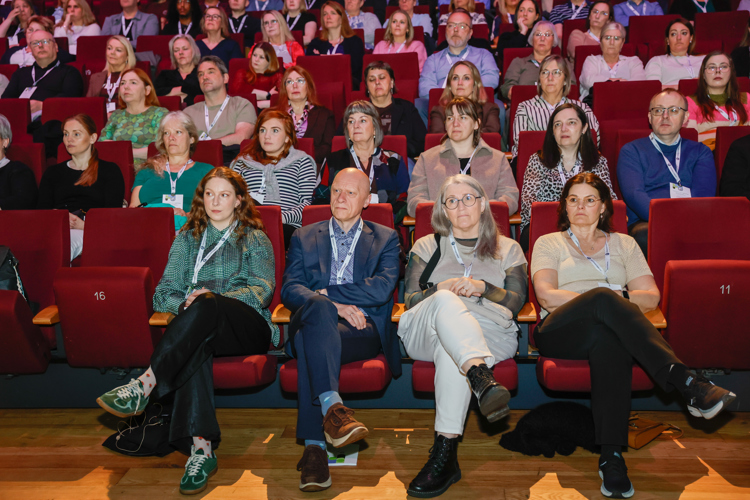 A diverse audience seated in a theater, attentively watching an event. Red seats and a wooden floor are visible.