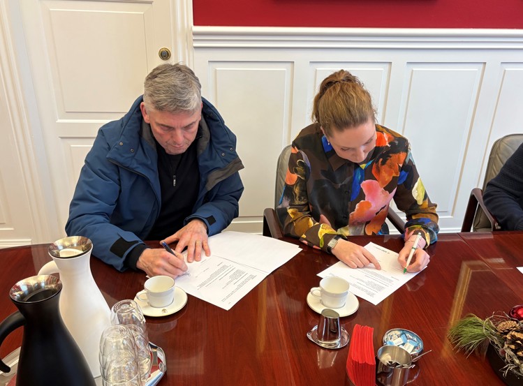 Two individuals are seated at a table, signing documents. A coffee service and decorative items are visible on the table.