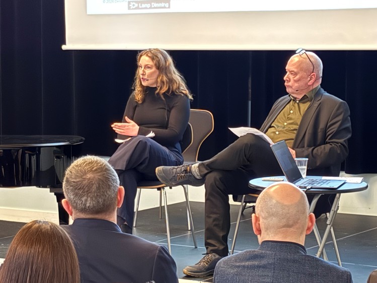 A panel discussion featuring two speakers, one gesturing while speaking, seated in front of an audience. A laptop and glass of water are on the table.