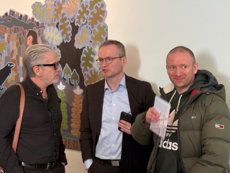 Three men are engaged in conversation, with a colorful mural featuring trees and figures in the background.
